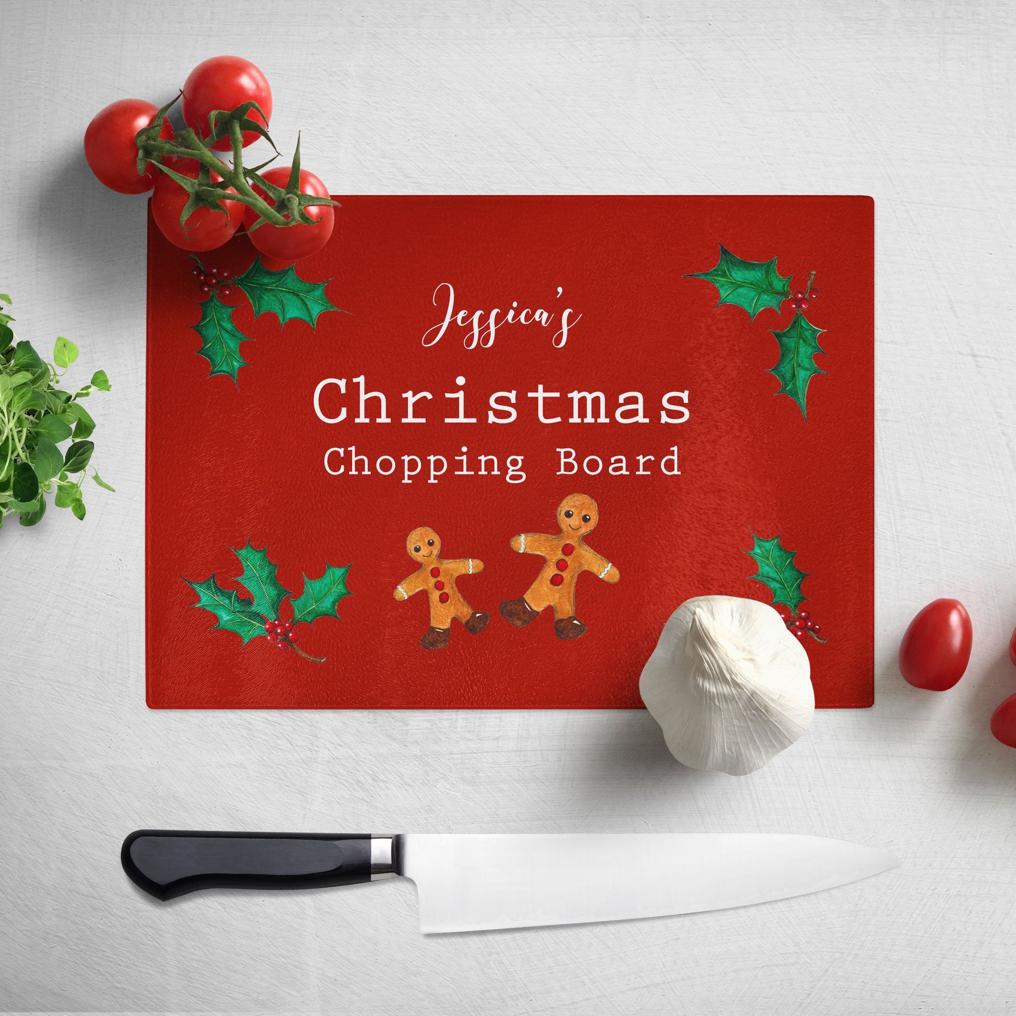 A personalised Christmas chopping board. The chopping board has a red background and a pattern of gingerbread men and holly leaves. The design includes white text which reads "Jessica's Christmas chopping board"