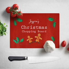 A personalised Christmas chopping board. The chopping board has a red background and a pattern of gingerbread men and holly leaves. The design includes white text which reads "Jessica's Christmas chopping board"