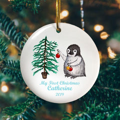 A personalised Christmas bauble with a penguin and a Christmas tree printed on it along with the message "My first Christmas, Catherine 2019"