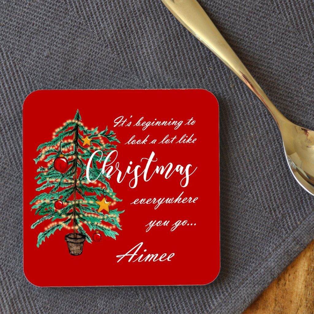 A personalised red square coaster with an illustration of a Christmas tree and the words "it's beginning to look a lot like Chrsitmas" printed onto it. The coaster is customised with the name "Aimee"
