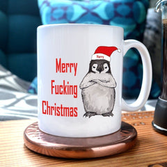 A personalised Christmas mug with the words "merry fucking Christmas" next to an illustration of a grumpy penguin in a Santa hat