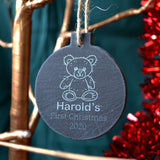A personalised round slate christmas bauble with an engraved design. The design features a teddy bear icon at the top and the words "Harold's first Christmas 2020" below.