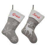 2 Christmas stockings in pale grey and white, one has a snowflake pattern and one has a stylised reindeer. Both are embroidered with a name in red on the white top of the stocking.